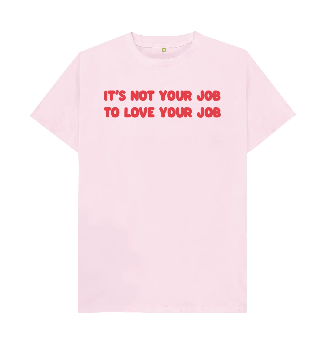 A light pink t-shirt with a large dark pink text along the front on two lines reading It's Not Your Job To Love Your Job.